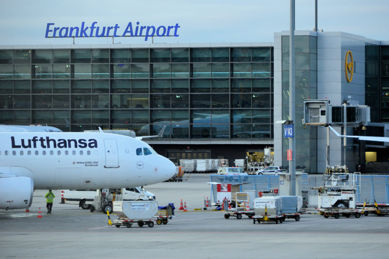 A Lufthansa aircraft at Frankfurt Airport with the modern terminal and bustling cargo operations in the background.