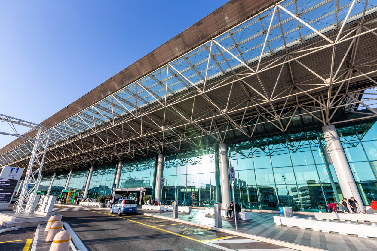 The glass-fronted Terminal 1 of Rome Fiumicino Airport displays modern architecture and a vibrant, welcoming entrance under a bright sky.