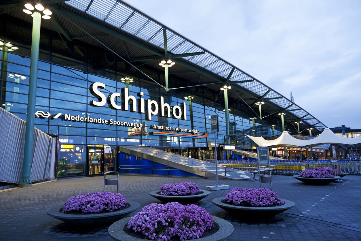 The illuminated entrance of Amsterdam Airport Schiphol, featuring its iconic signage and vibrant flower displays under a twilight sky.