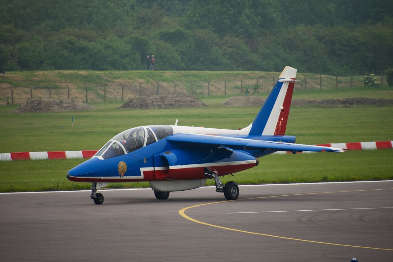 A vibrant aircraft is seen taxiing on the runway at Cambridge Airport, surrounded by green fields and a clear backdrop.