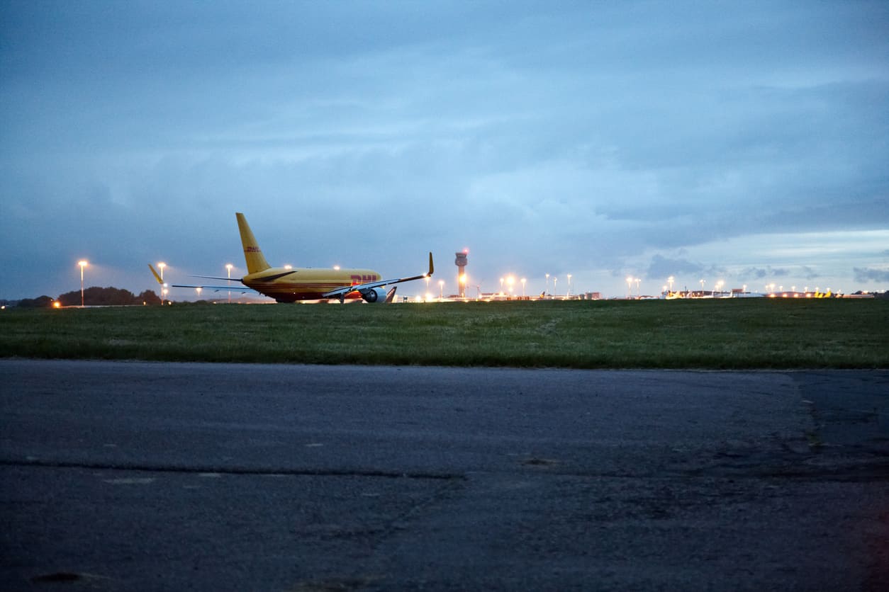 A DHL cargo plane rests on the runway at East Midlands Airport, with the airport's control tower glowing in the evening light.