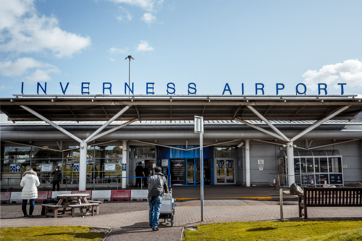 The main entrance of Inverness Airport, featuring its bold signage and a few travelers approaching the terminal under a bright, partly cloudy sky.