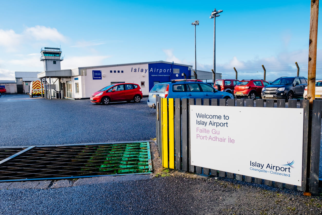 Islay Airport's entrance and terminal on a sunny day, with a welcoming sign and parked vehicles in view.