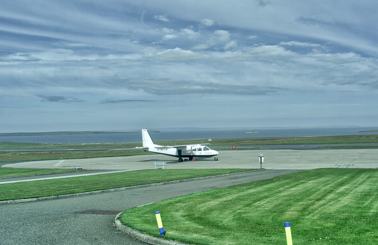 A small aircraft on the tarmac at Kirkwall Airport, with the serene Orkney Islands and sea in the background.