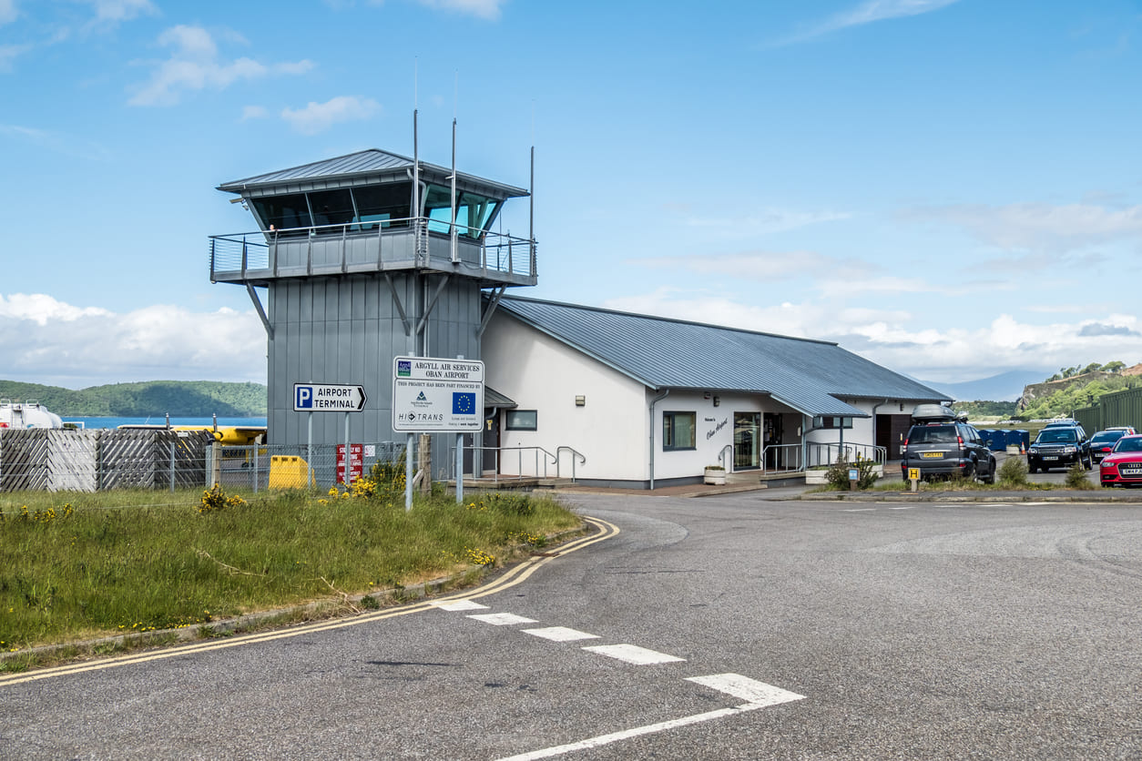 The terminal and control tower of Oban Airport, nestled amidst scenic landscapes under a clear blue sky.