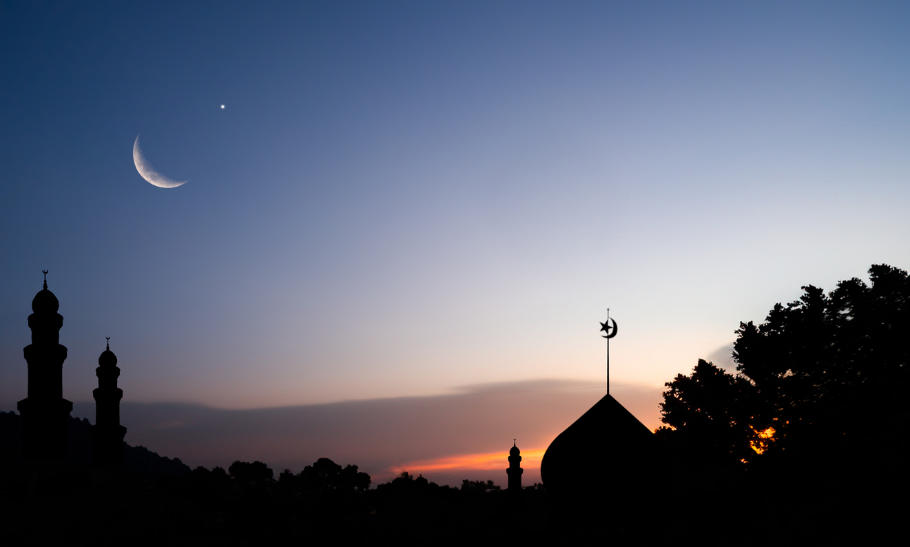 Crescent moon above mosque silhouettes at dusk, symbolizing the start of a new month in the Islamic calendar.