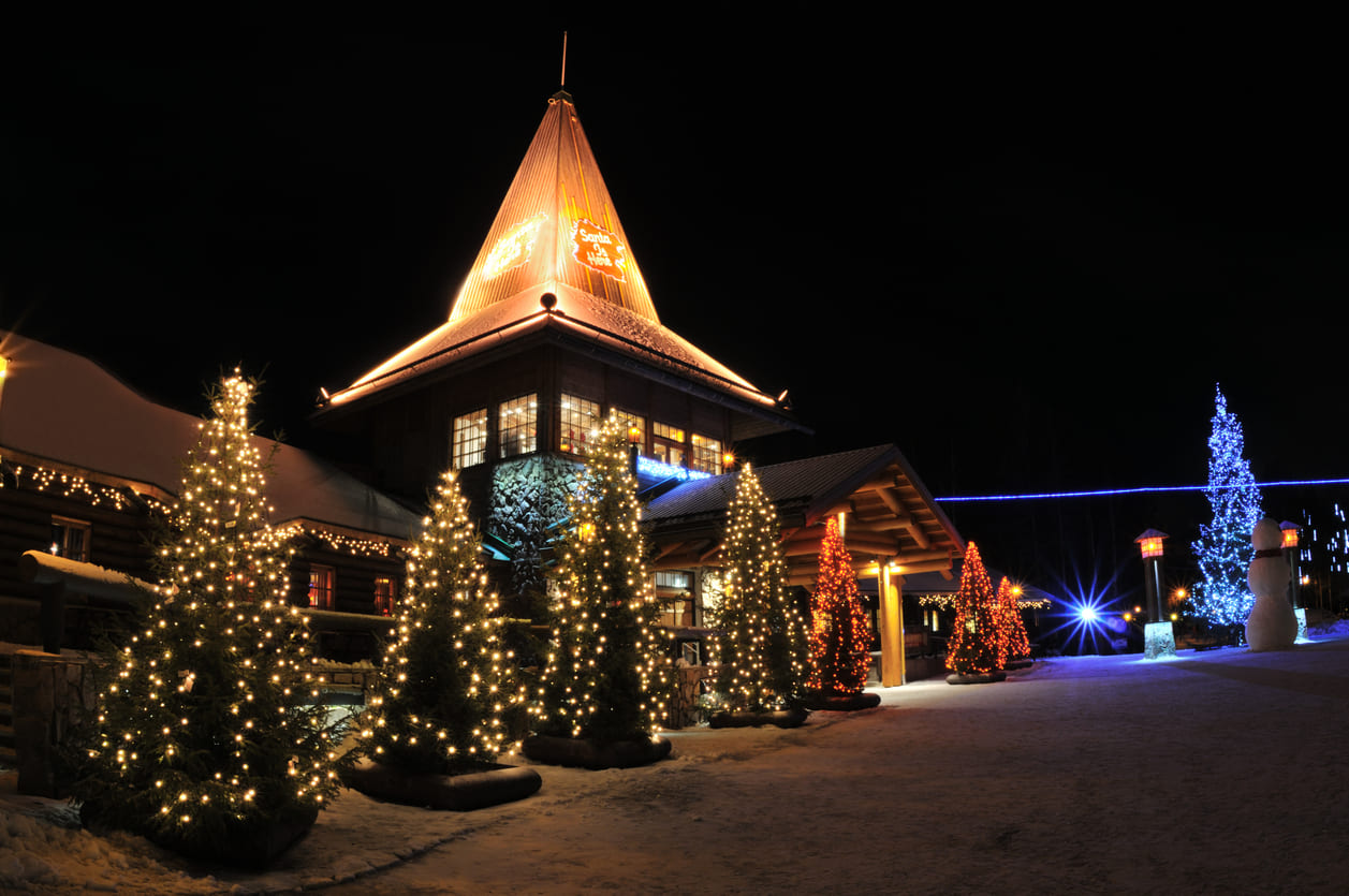 A snowy evening at Santa Claus Village in Rovaniemi, illuminated by glowing Christmas trees and festive holiday lights against the dark Arctic sky.