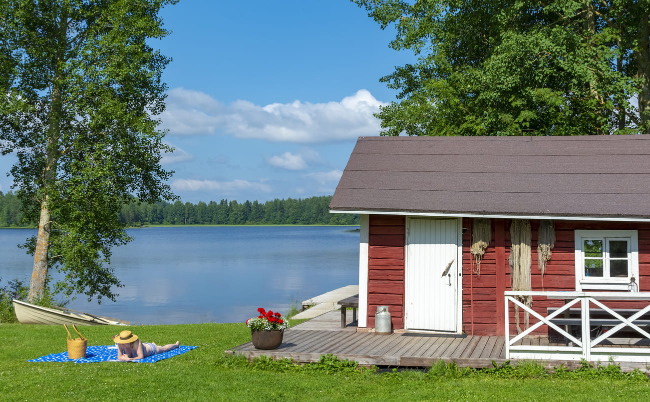 A tranquil Finnish lakeside with a red summer cottage and a person relaxing on a blanket, reflecting the calm and natural beauty of Midsummer Day.