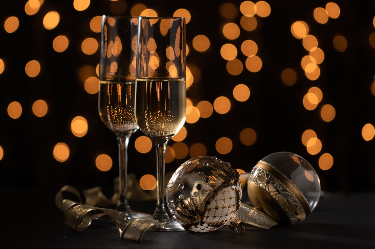 Two champagne glasses and elegant ornaments sit against a backdrop of glowing lights, evoking the festive mood of New Year's Eve.