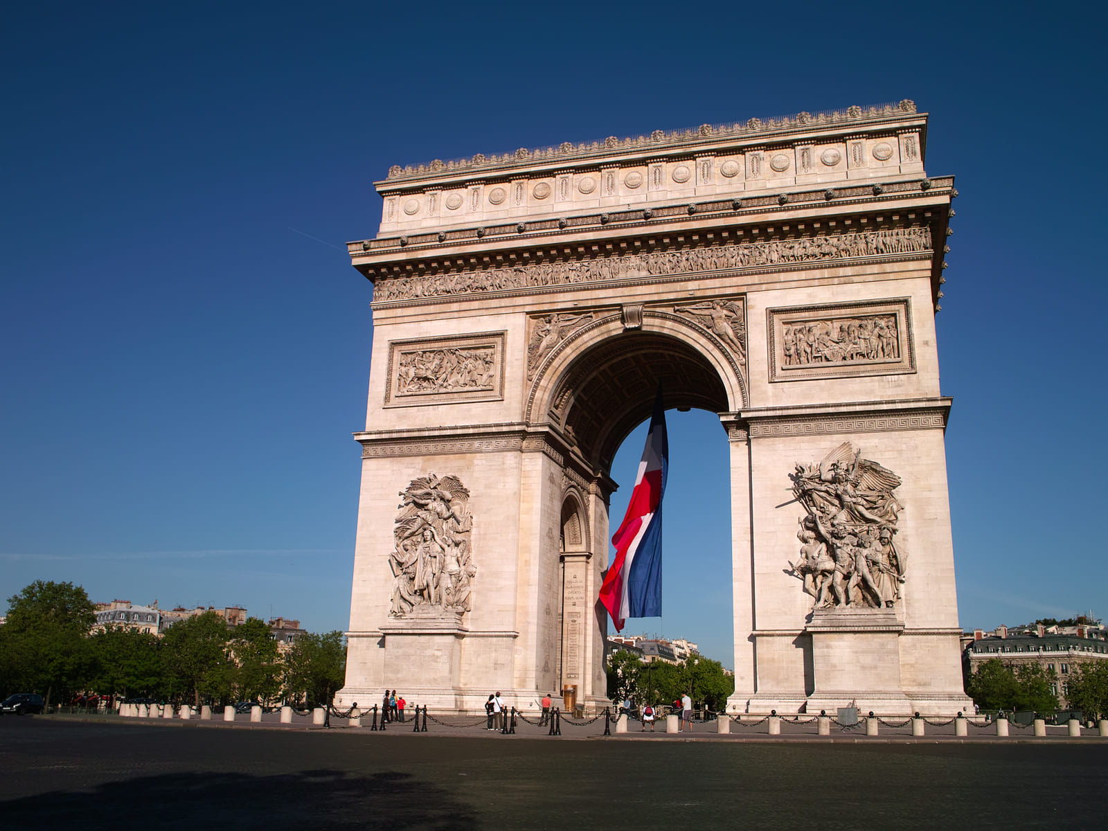 The Arc de Triomphe stands under a clear blue sky with a large French flag hanging in its central arch.