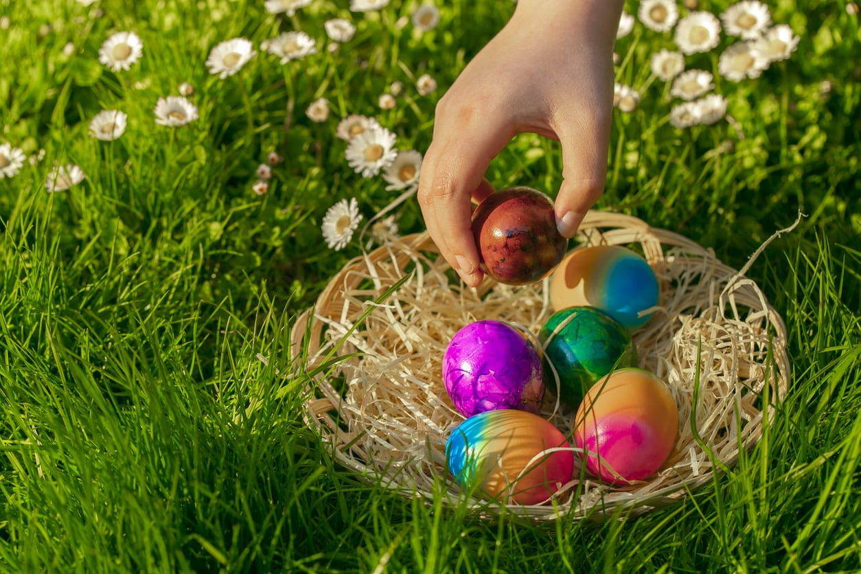 A hand reaches into a basket of vibrant Easter eggs resting on lush green grass, capturing the spirit of an Easter egg hunt.