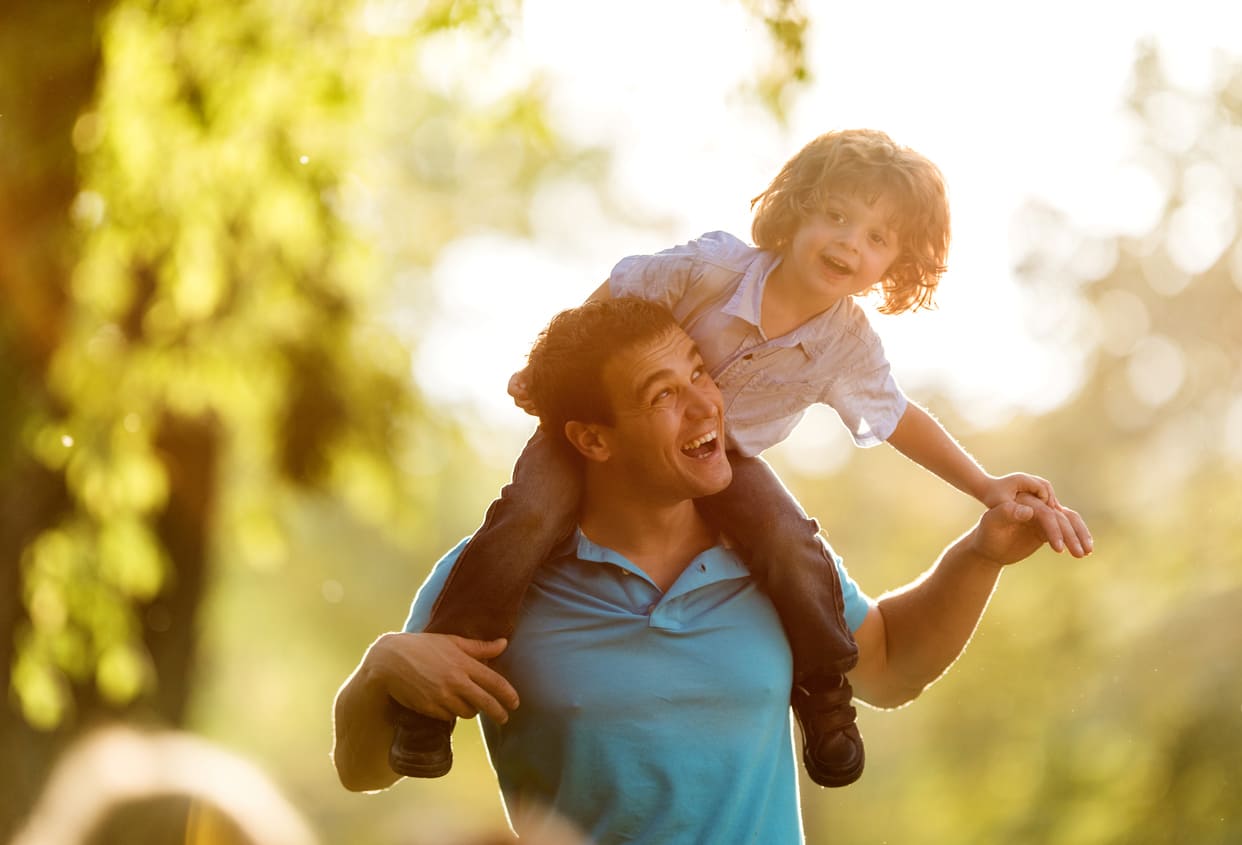 A father smiles as he carries his happy child on his shoulders, enjoying a sunny day together.