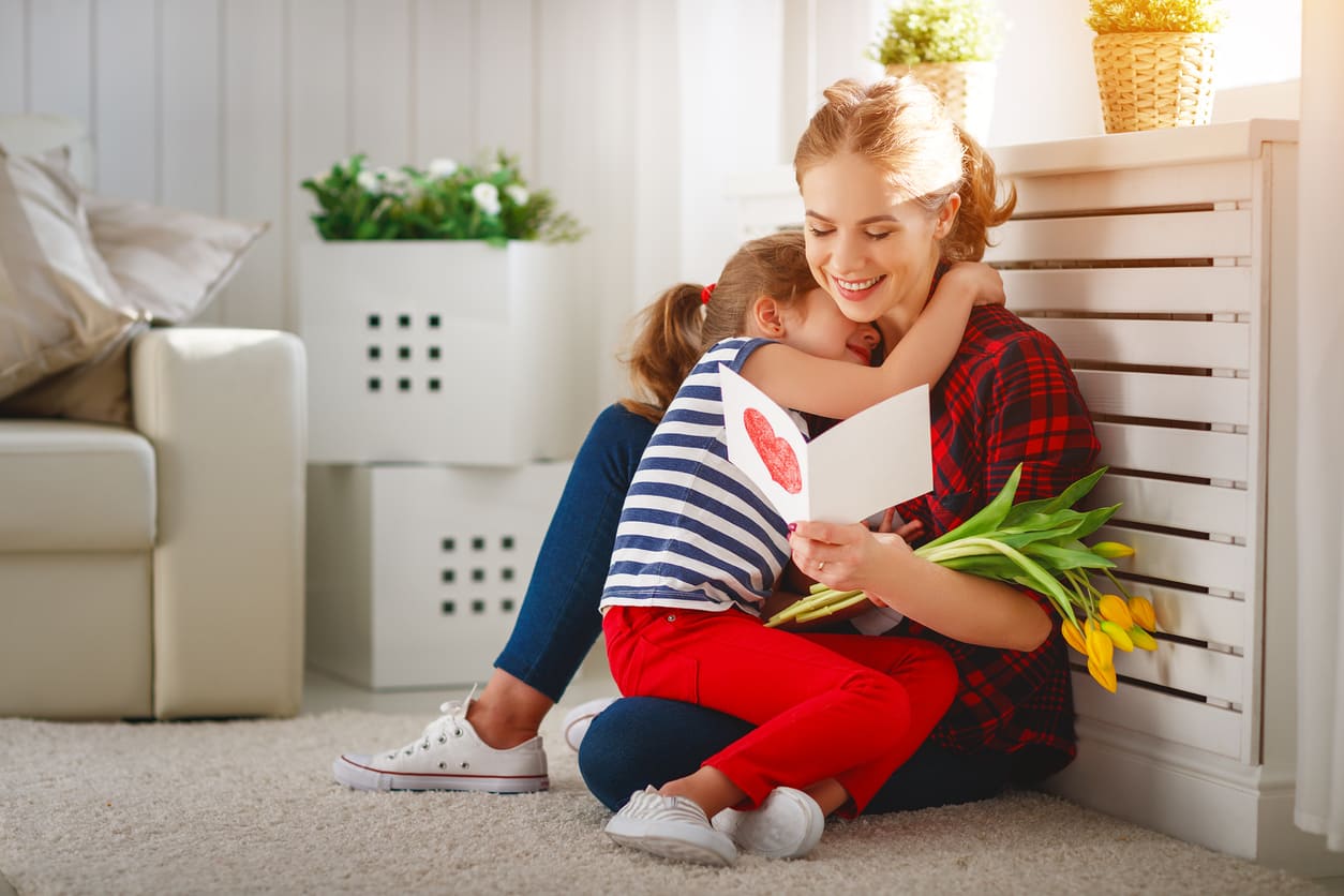 A mother and child share a warm embrace as the child presents a handmade card and flowers, celebrating Mother's Day.