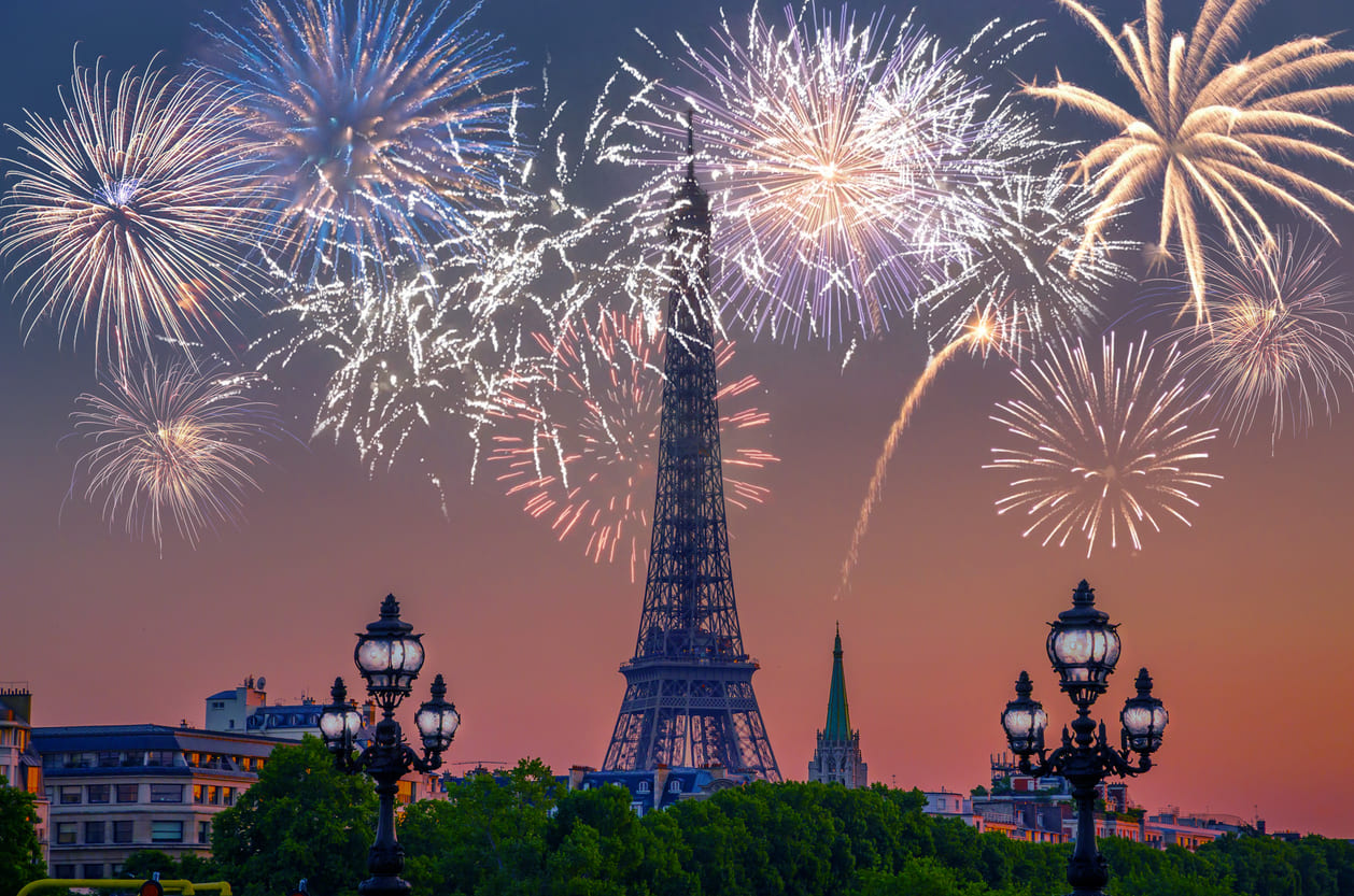 The Eiffel Tower stands illuminated against a twilight sky, surrounded by vibrant fireworks bursting in celebration.