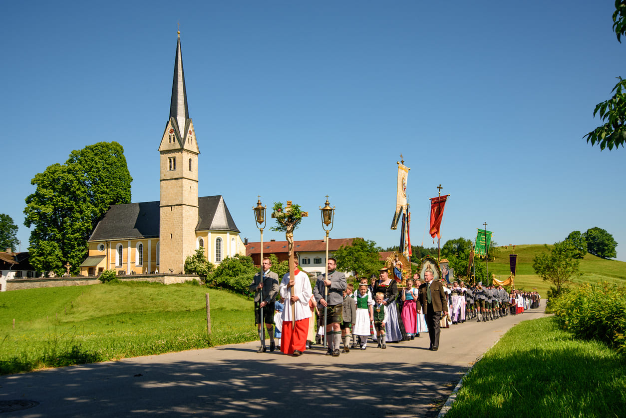 A traditional Corpus Christi procession in Germany, with participants carrying banners and crosses near a picturesque church.