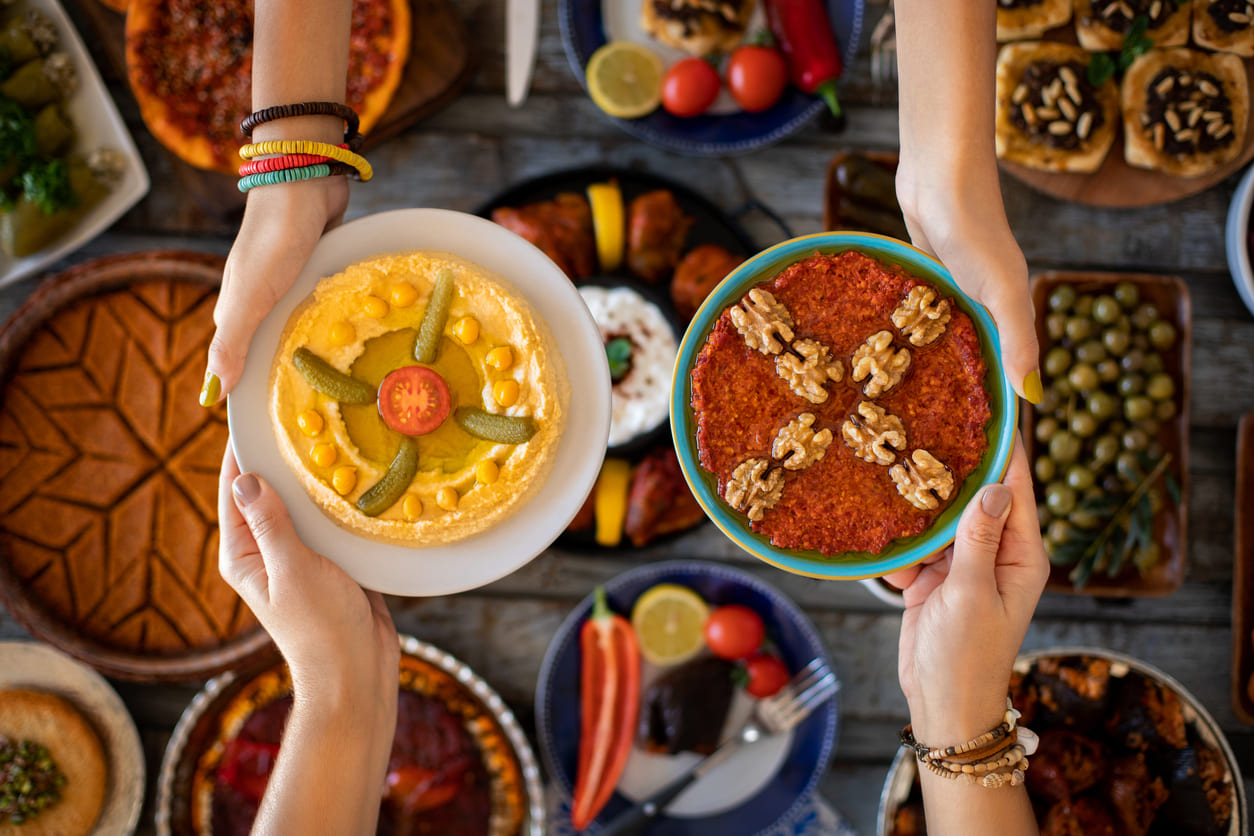 Hands holding plates of vibrant traditional dishes, surrounded by a festive table filled with food, reflecting the celebratory spirit of Eid al-Fitr.