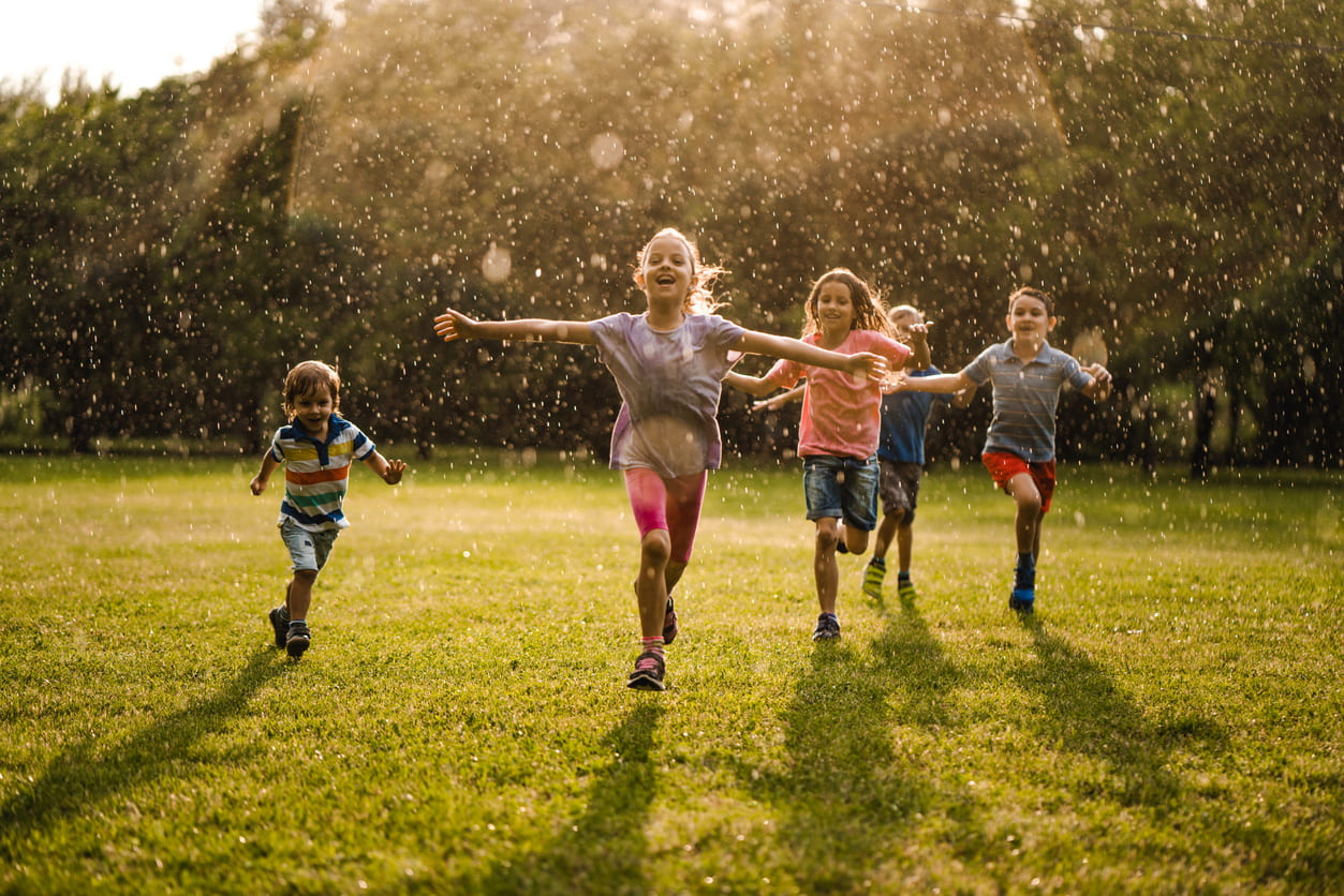 A group of joyful children running through a sunlit park, playing together in the refreshing drizzle.