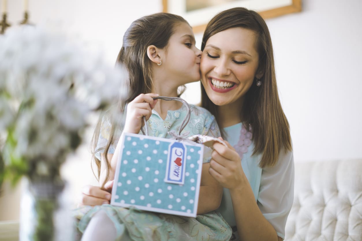 A young girl lovingly kisses her smiling mother while giving her a gift, capturing a warm Mother's Day moment.