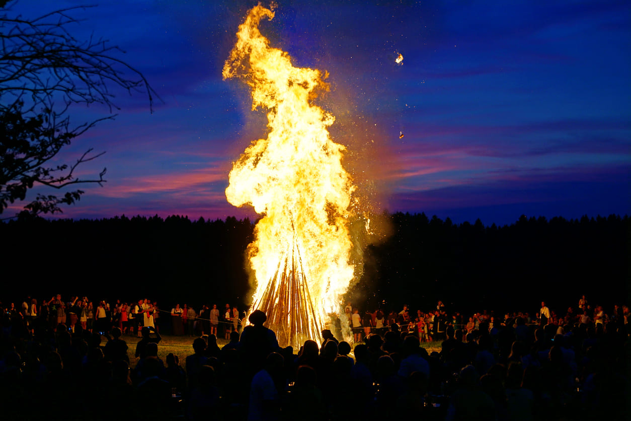 A towering Johannisfeuer burns brightly against the evening sky, with a crowd gathered to celebrate the traditions of St. John's Day.