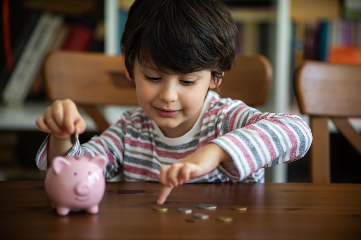 A young boy is carefully placing coins into a pink piggy bank, representing the importance of saving.