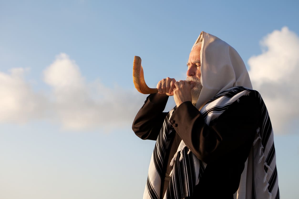 A man wrapped in a prayer shawl blowing a shofar, a key ritual symbolizing the conclusion of Yom Kippur and spiritual renewal.