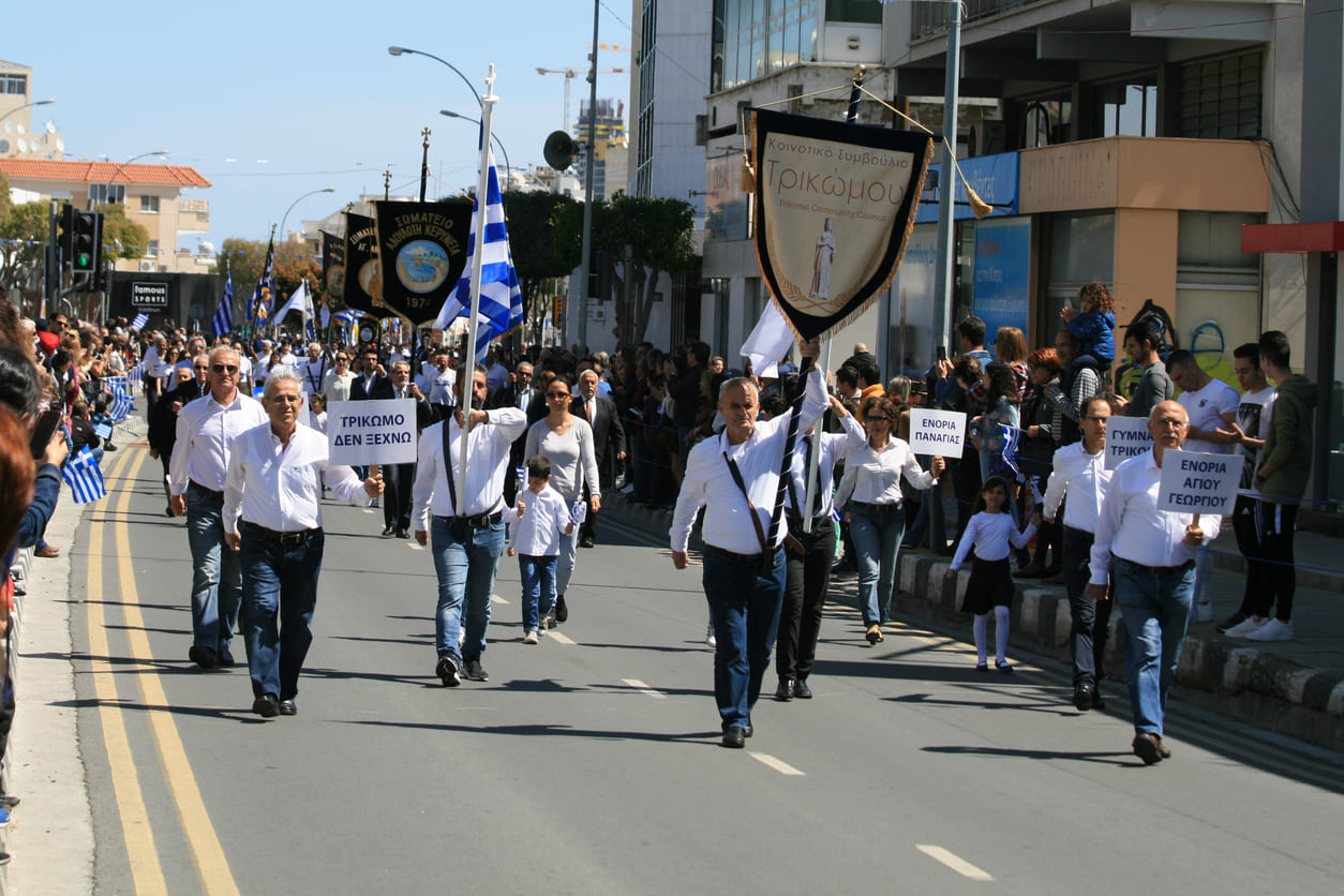 A community parade featuring Greek flags and banners fills the street as people of all ages commemorate Greek Independence Day with pride and unity.
