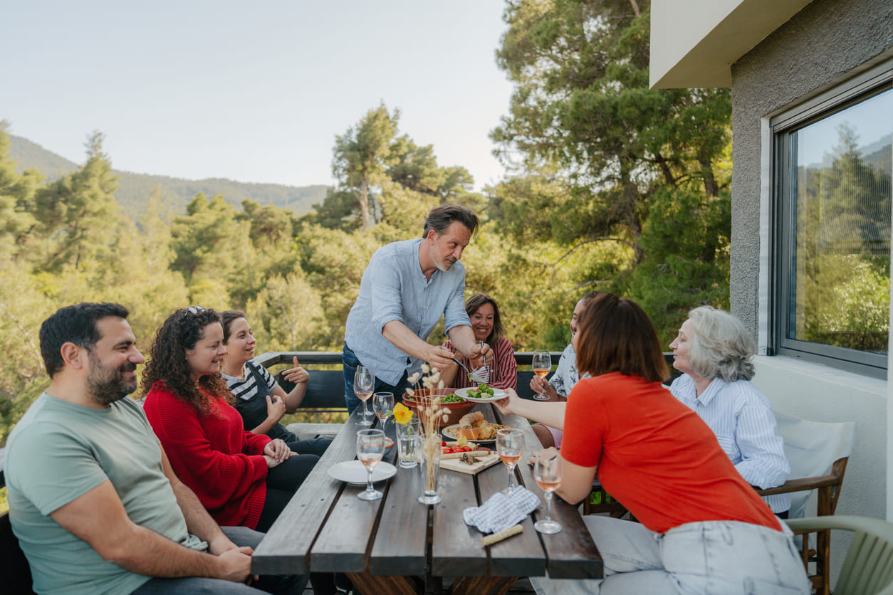 A cheerful Greek family gathers around a wooden table on a sunny terrace, sharing food and laughter on Orthodox Easter Monday.