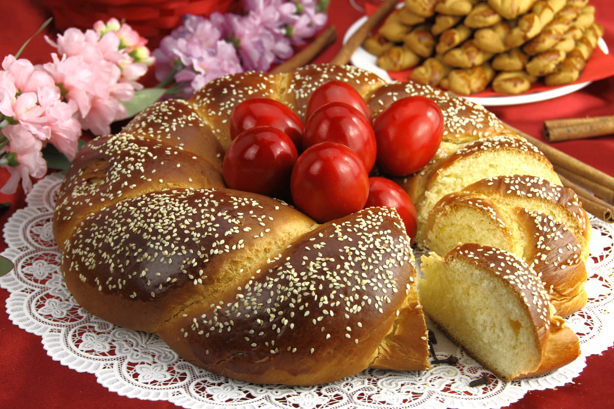 A beautifully braided tsoureki loaf topped with sesame seeds surrounds vibrant red-dyed eggs, creating a traditional and symbolic centerpiece for Greek Orthodox Easter.