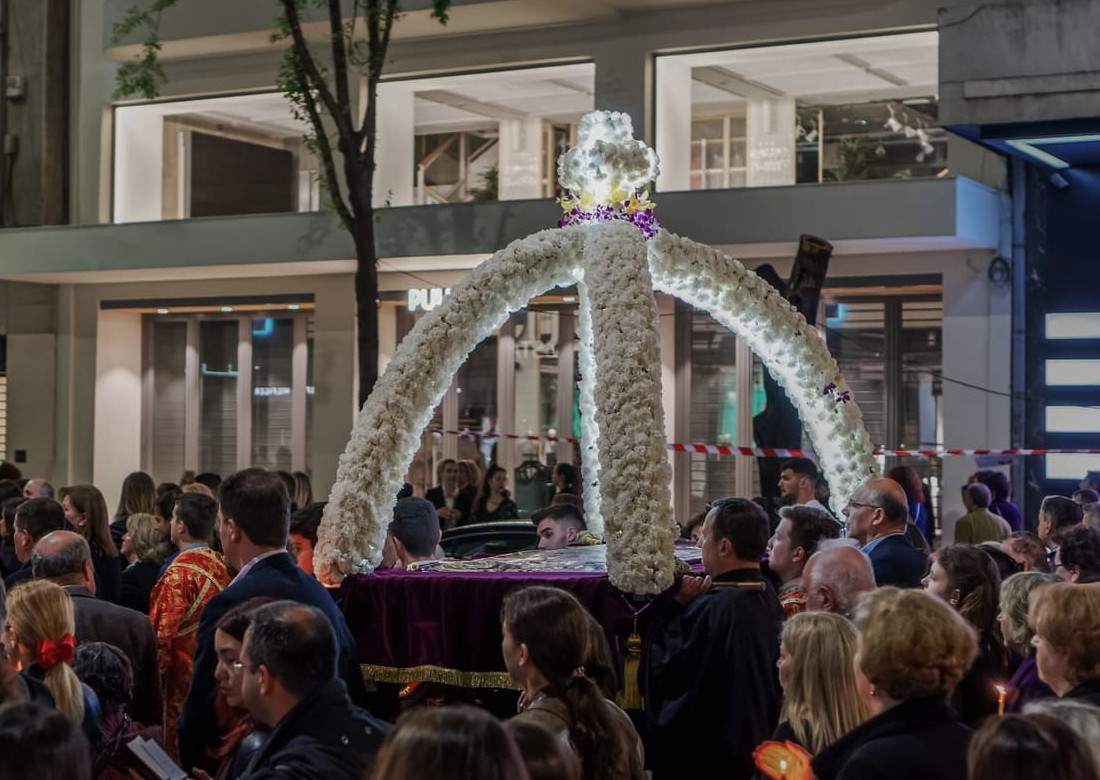 A crowd of Greek worshippers walks reverently through city streets at night, following a flower-covered Epitaphios during the Orthodox Good Friday procession.