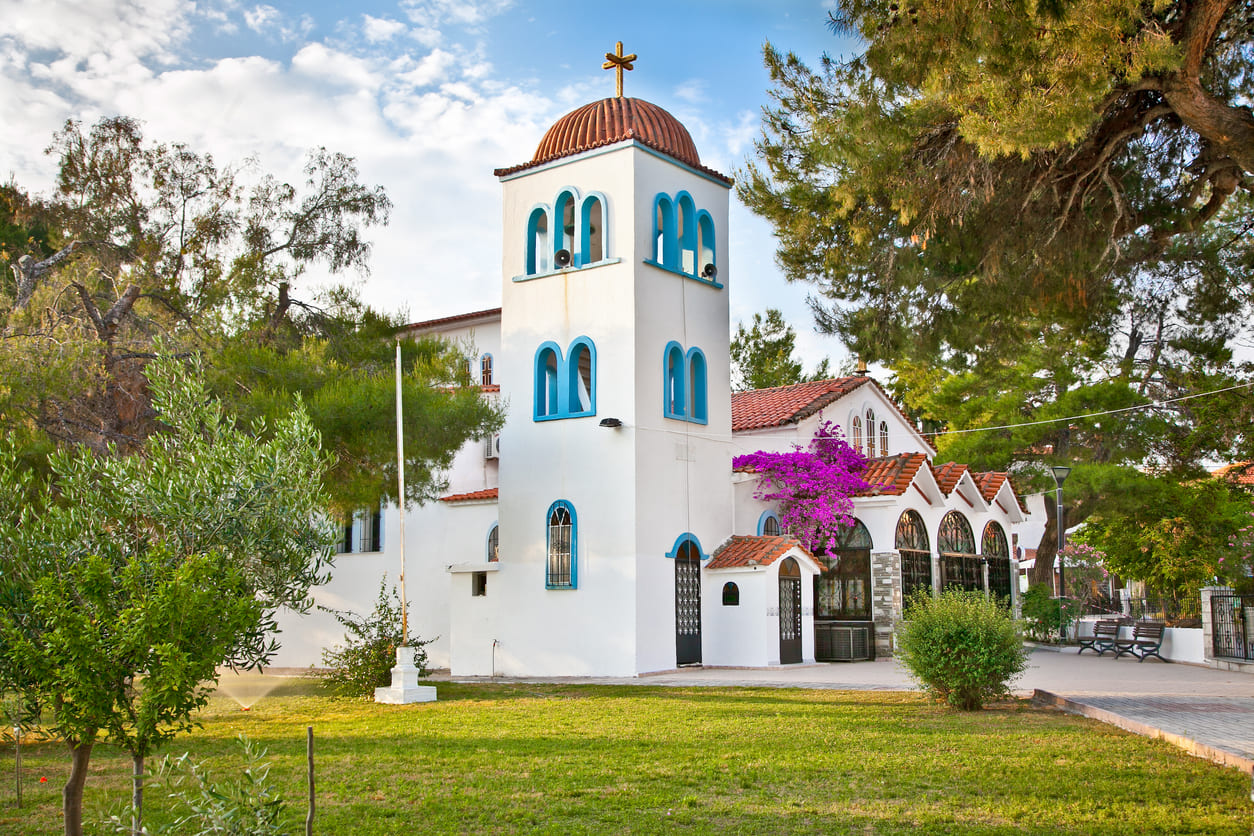 A whitewashed Orthodox church with blue accents and blooming flowers stands peacefully amid trees on the Kassandra Peninsula, capturing the calm of Holy Spirit Monday.
