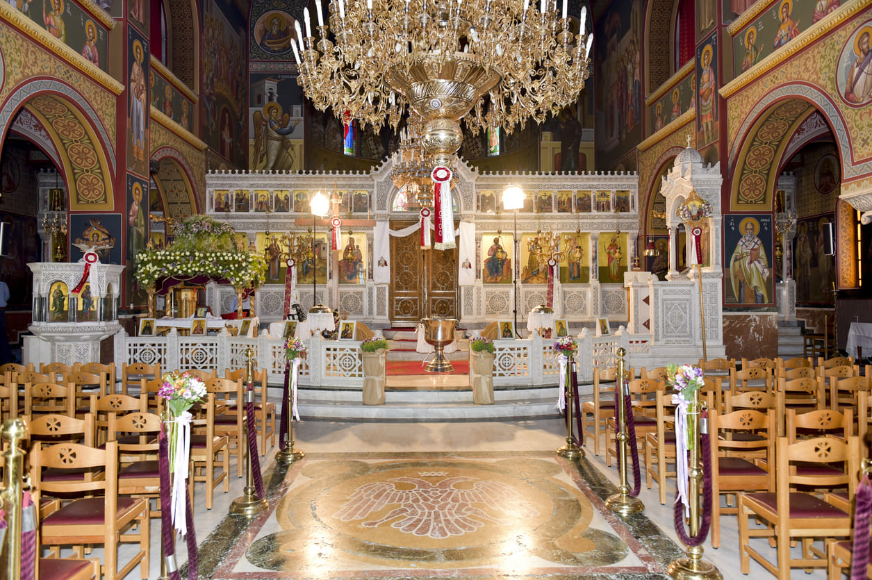 An ornately decorated Greek Orthodox church interior, with chandeliers, icons, and floral arrangements, set for the Pentecost celebration.
