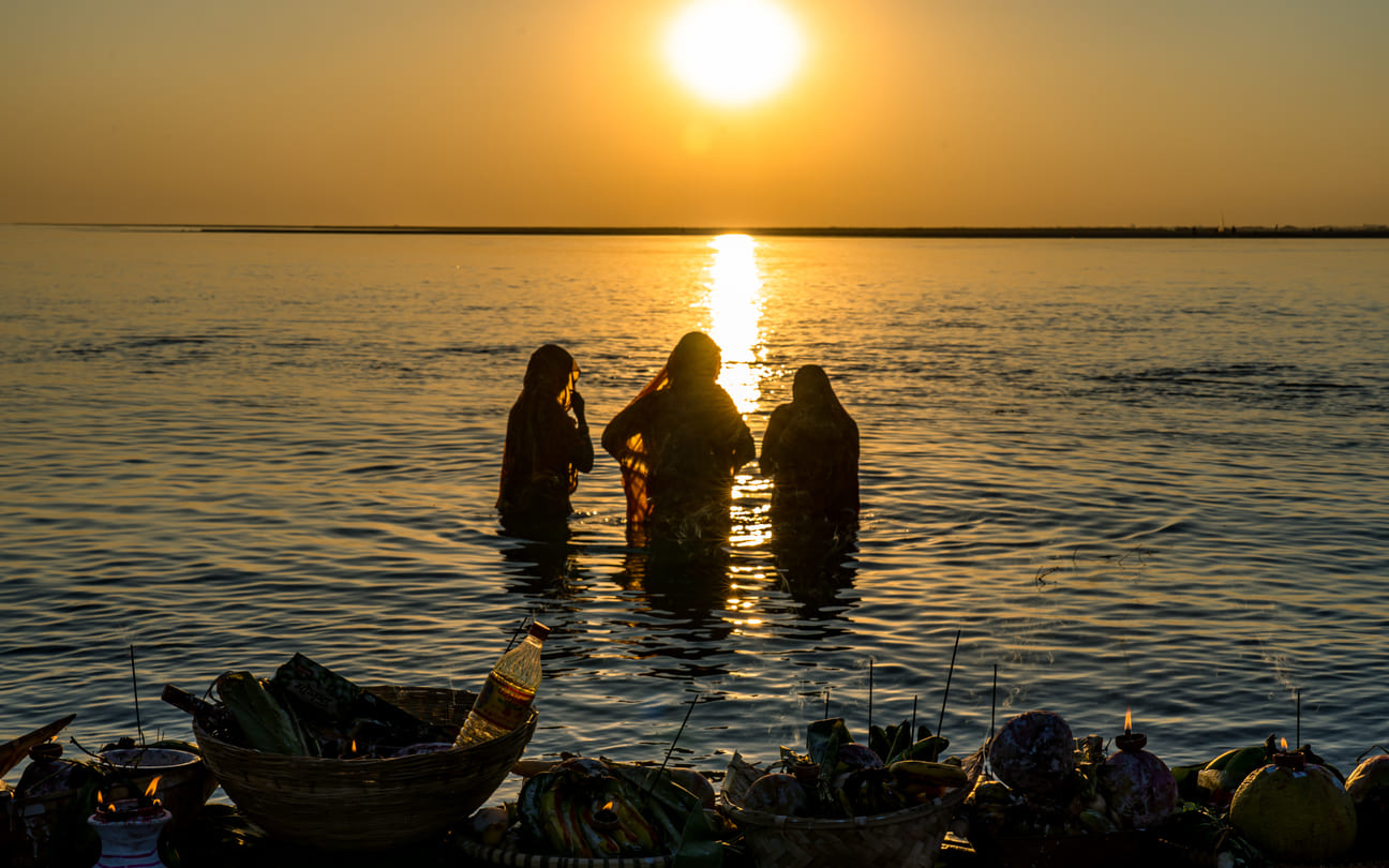 Women standing in a river during sunrise, performing Chhath Puja rituals with offerings placed nearby.