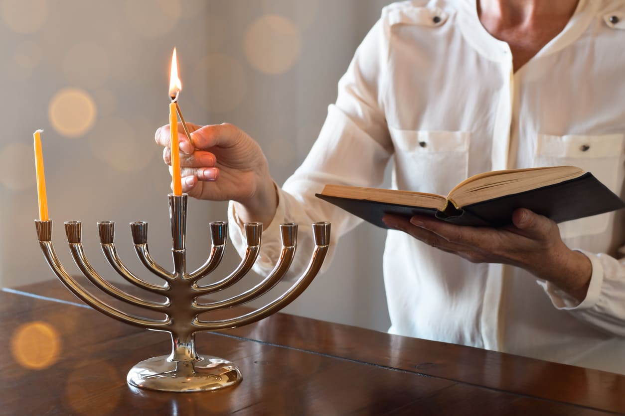 A person lights the menorah while holding a prayer book, marking the beginning of Hanukkah.