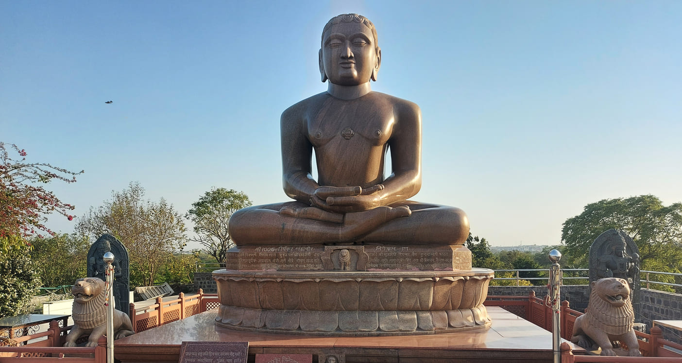 Majestic statue of Lord Mahavira seated in meditation, exuding peace and spirituality amidst a tranquil outdoor setting.