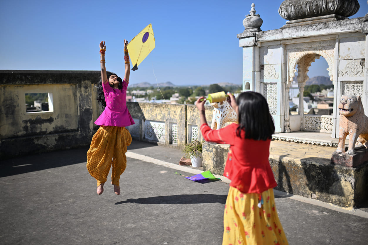 Two children are joyfully flying kites on a rooftop, capturing the festive essence of Makar Sankranti under a bright blue sky.