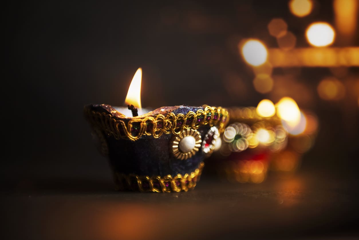 A decorated clay oil lamp (diya) glows warmly in the dark, symbolizing the light and hope of Deepavali amid a backdrop of soft, festive lights.