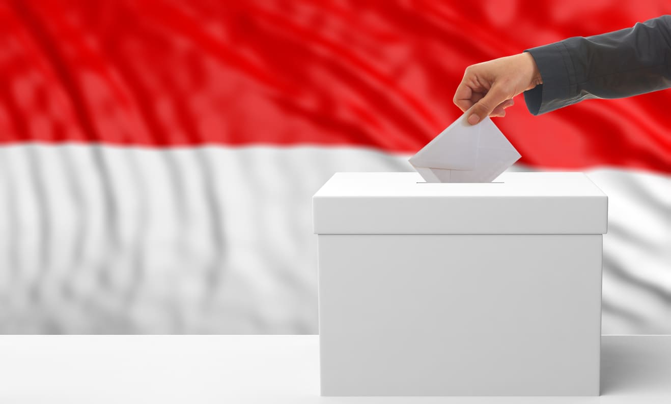A close-up of a hand placing a ballot into a box with the Indonesian flag in the background, symbolizing the act of voting.