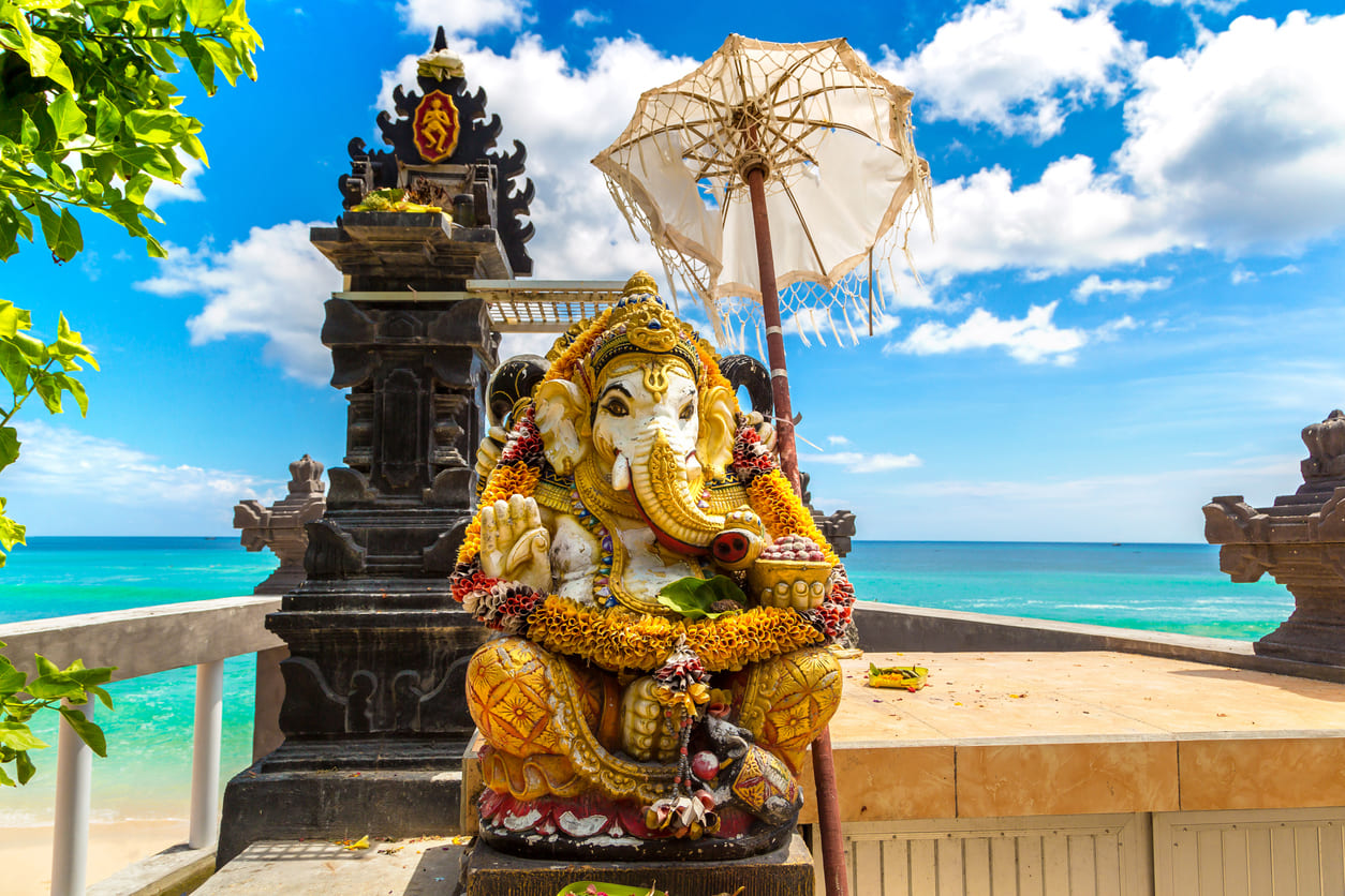 A vibrant statue of Lord Ganesha, adorned with flower garlands, stands at a beachside temple in Bali beneath a clear blue sky.