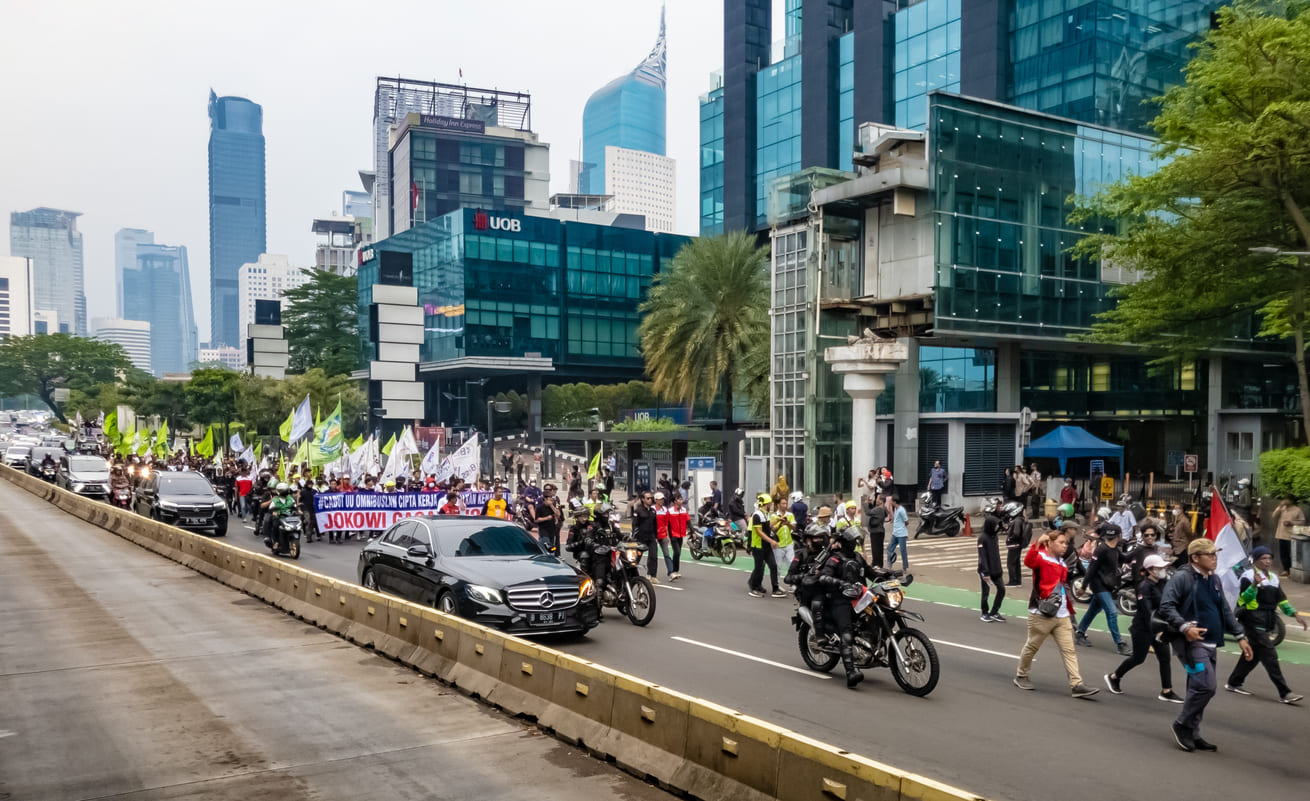 A vibrant street scene in Jakarta with workers marching alongside vehicles, holding banners and flags amid modern city buildings.
