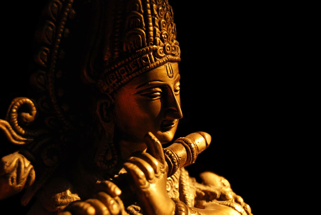 A detailed statue of Lord Krishna standing and playing the flute, adorned with flowers and traditional ornaments.