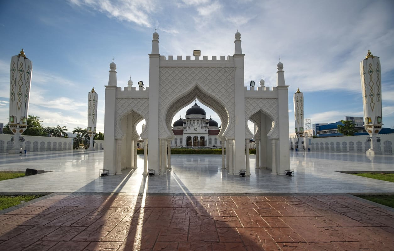 A beautiful view of the Baiturrahman Grand Mosque framed by its elegant white archways under a clear blue sky.