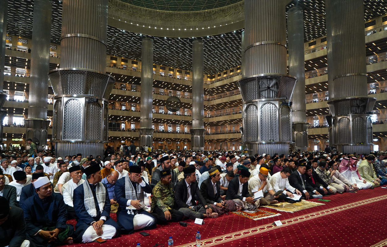 A wide view of worshippers seated inside the grand Istiqlal Mosque, surrounded by towering columns and intricate modern architecture.