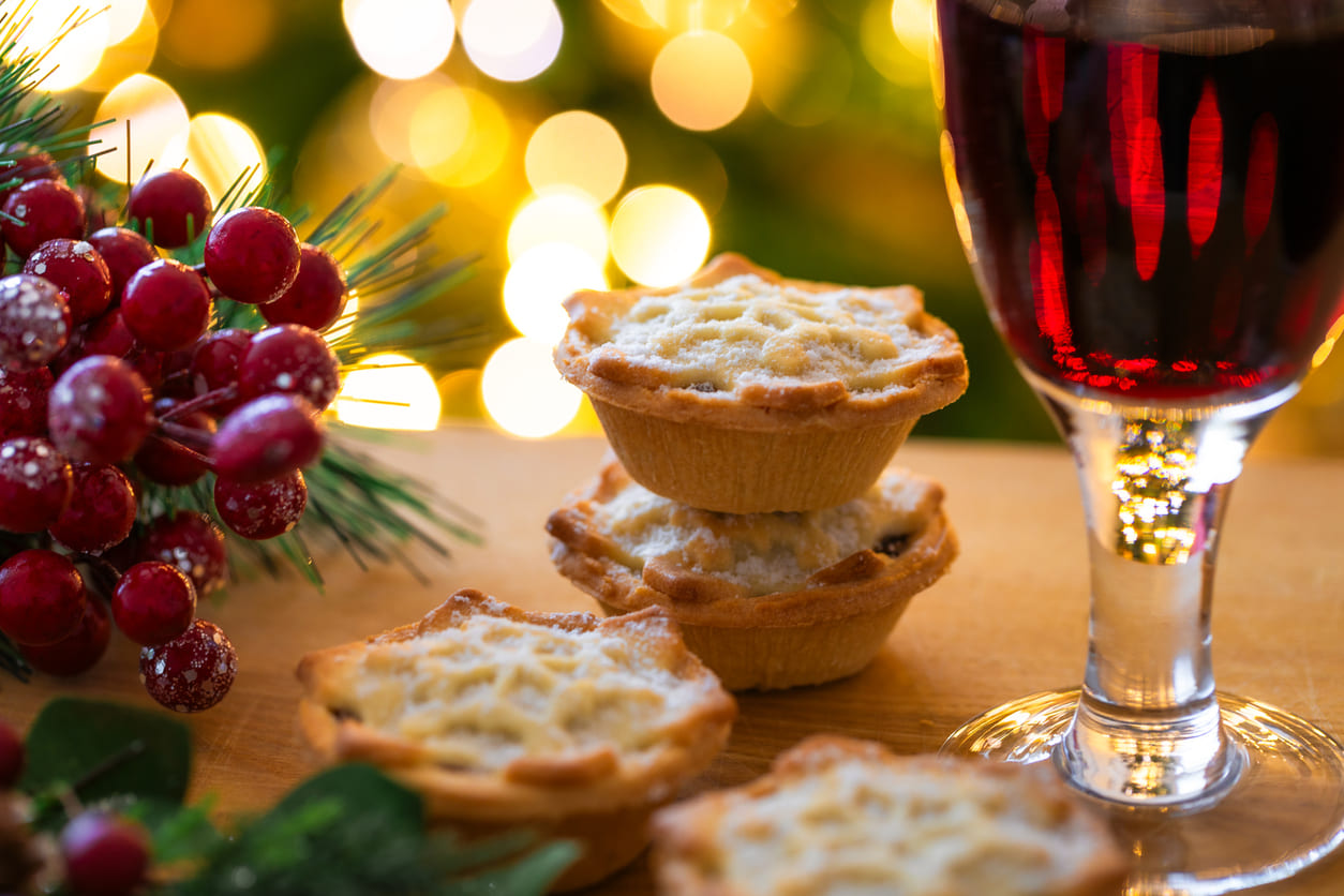 A festive scene featuring mince pies, a glass of wine, and holiday decorations glowing warmly in the background.