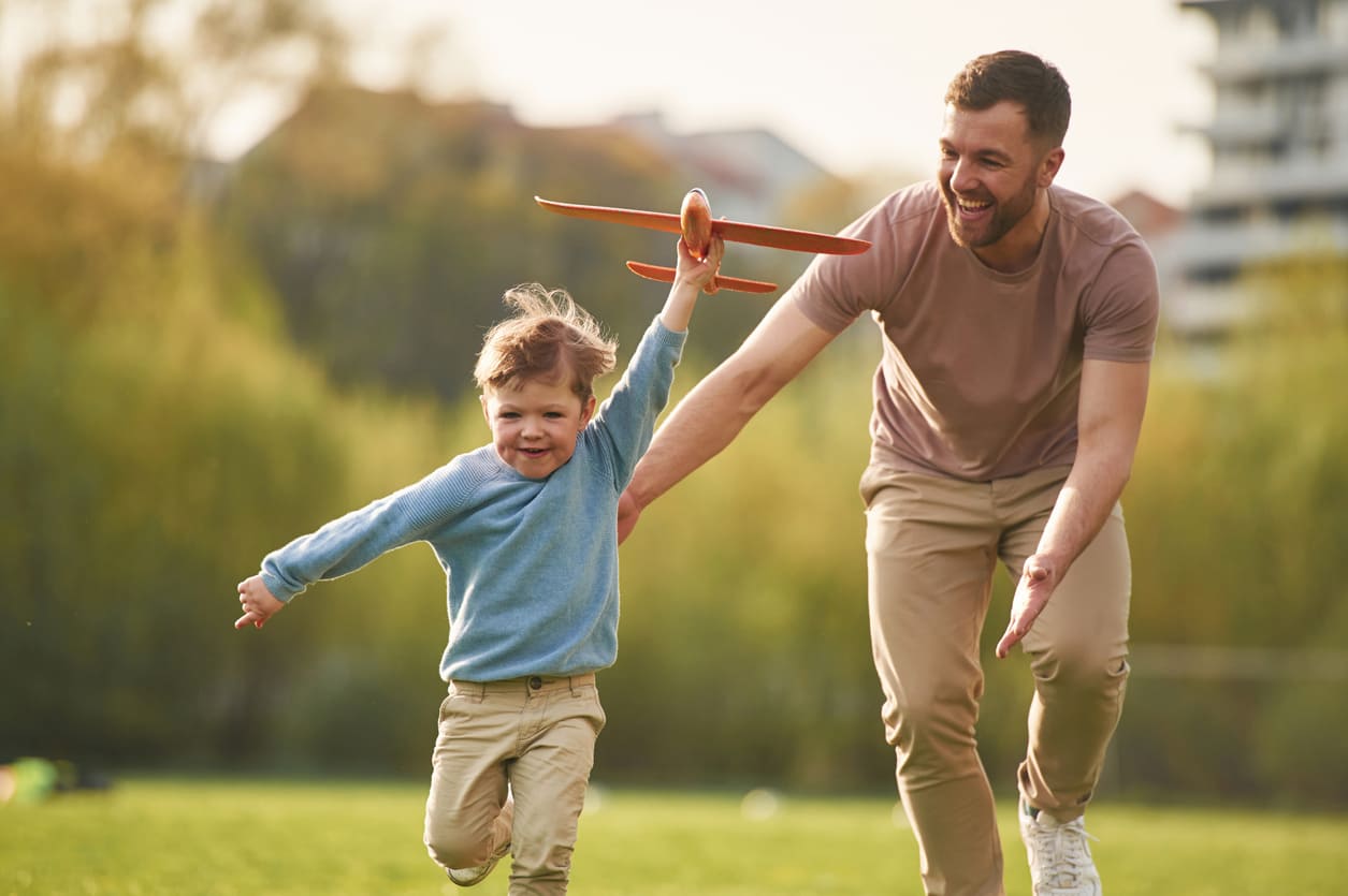 A father joyfully runs alongside his young son holding a toy airplane, sharing a playful and loving moment together outdoors.