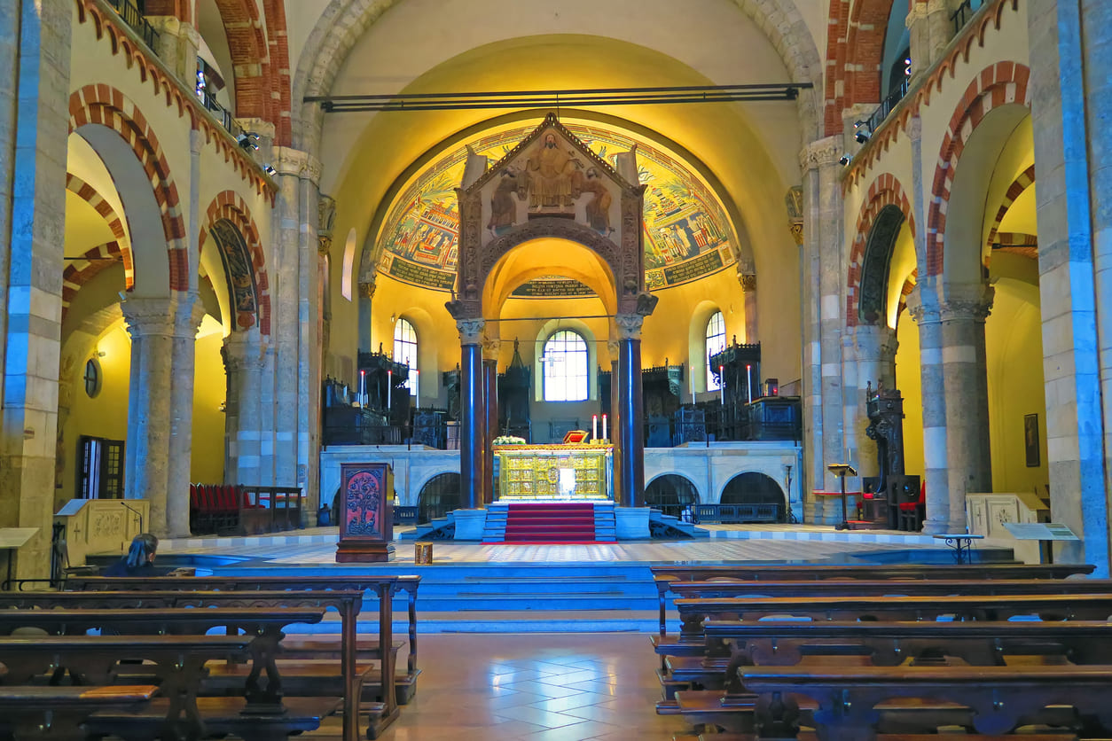 The Basilica of Sant'Ambrogio in Milan features a grand, illuminated interior with intricate arches and a central altar, capturing its historic beauty.