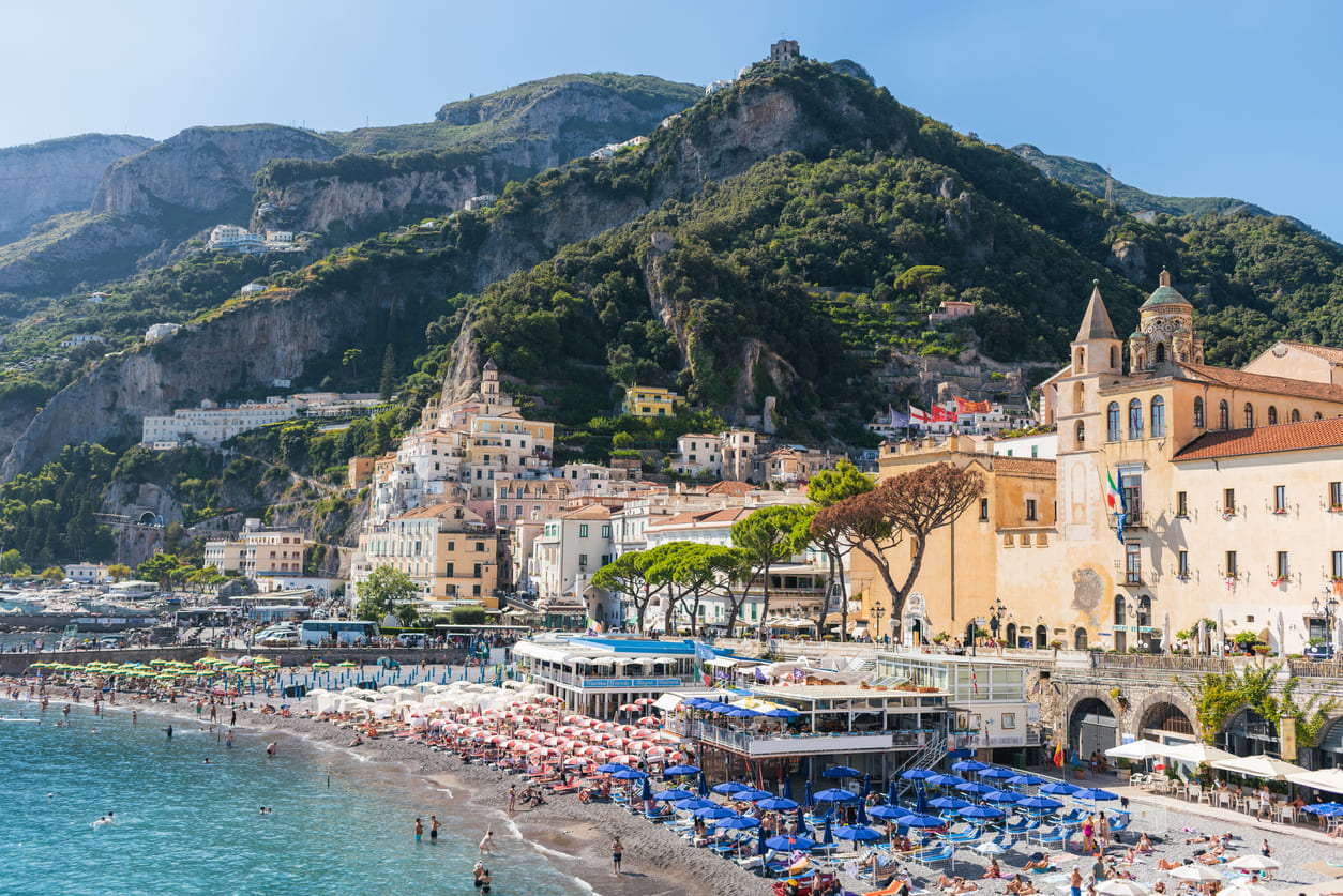 A lively beach scene on the Amalfi Coast, with colorful umbrellas and seaside buildings nestled against lush hills.