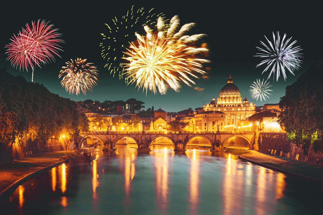 Fireworks illuminate the night sky over St. Peter's Basilica in Rome, with the river reflecting the vibrant colors.