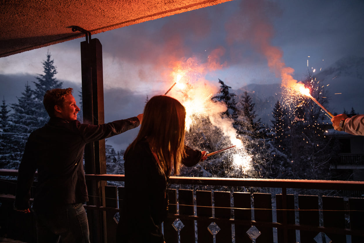 Three people hold sparklers on a balcony against a backdrop of snowy trees and a dusky sky.