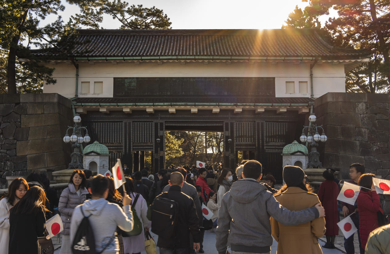 A crowd of people holding Japanese flags walks toward the Imperial Palace gate under warm sunlight, creating a festive and patriotic scene.
