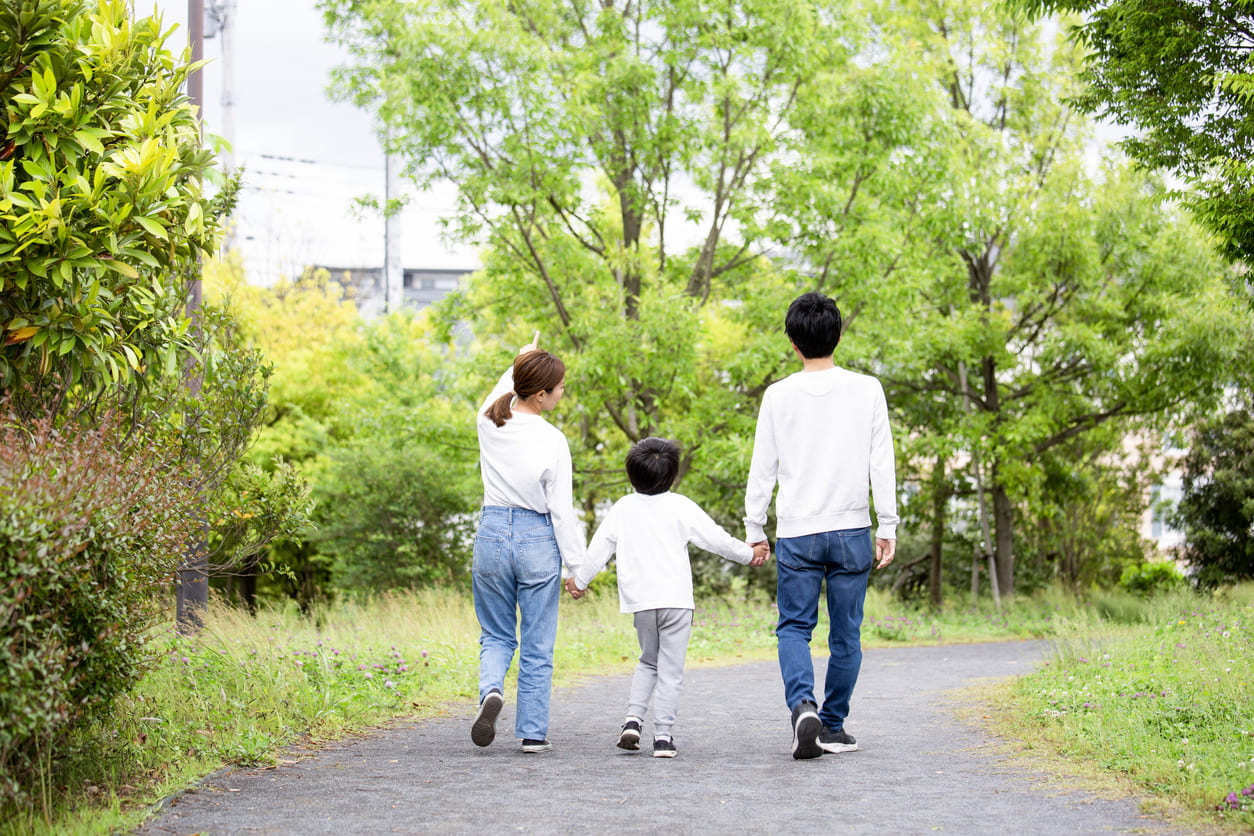 A family walks hand in hand along a tree-lined path, surrounded by lush greenery and blooming plants on a clear day.